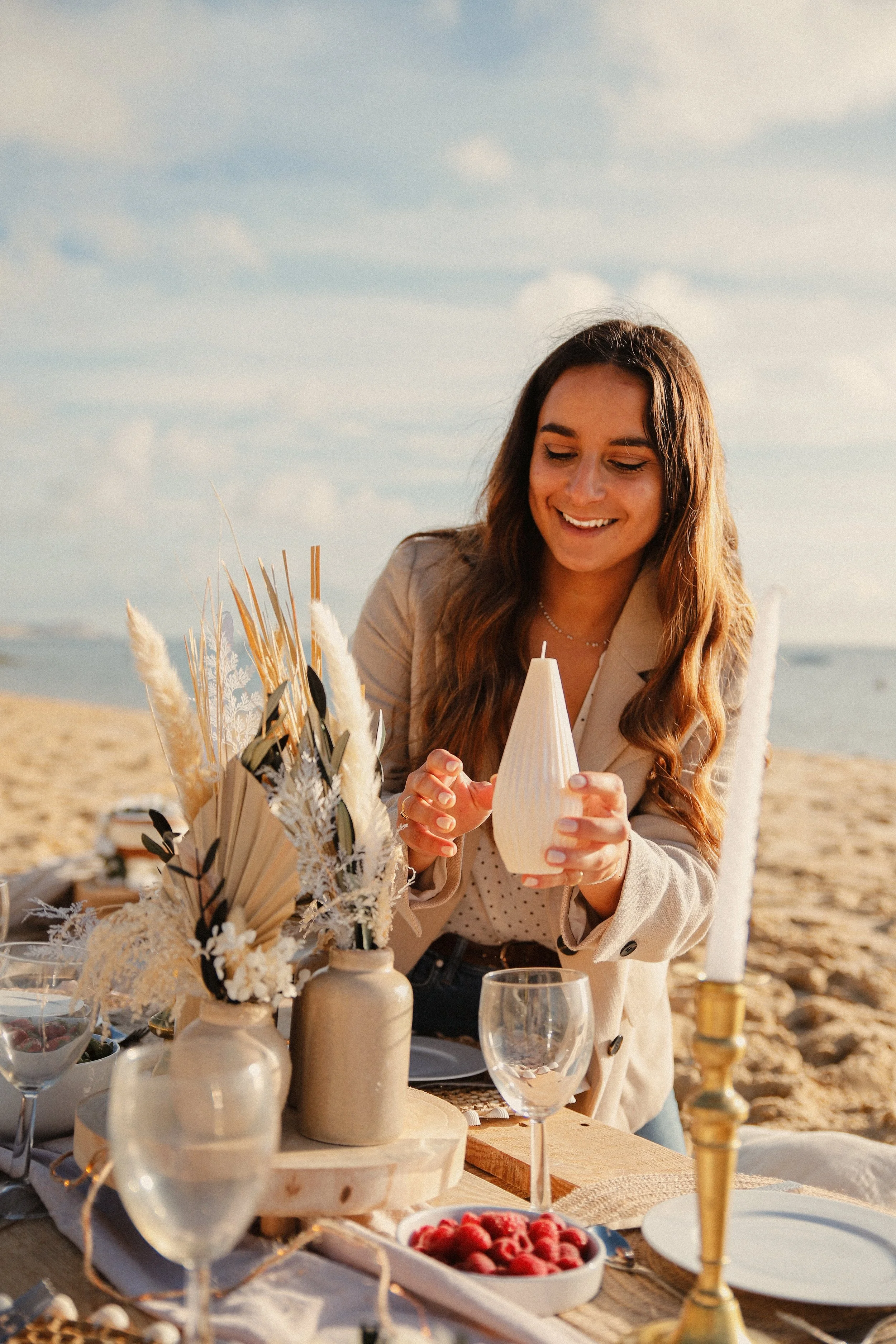 Femme souriante préparant une table de pique-nique sur la plage avec décoration florale, bougies et verre de vin.