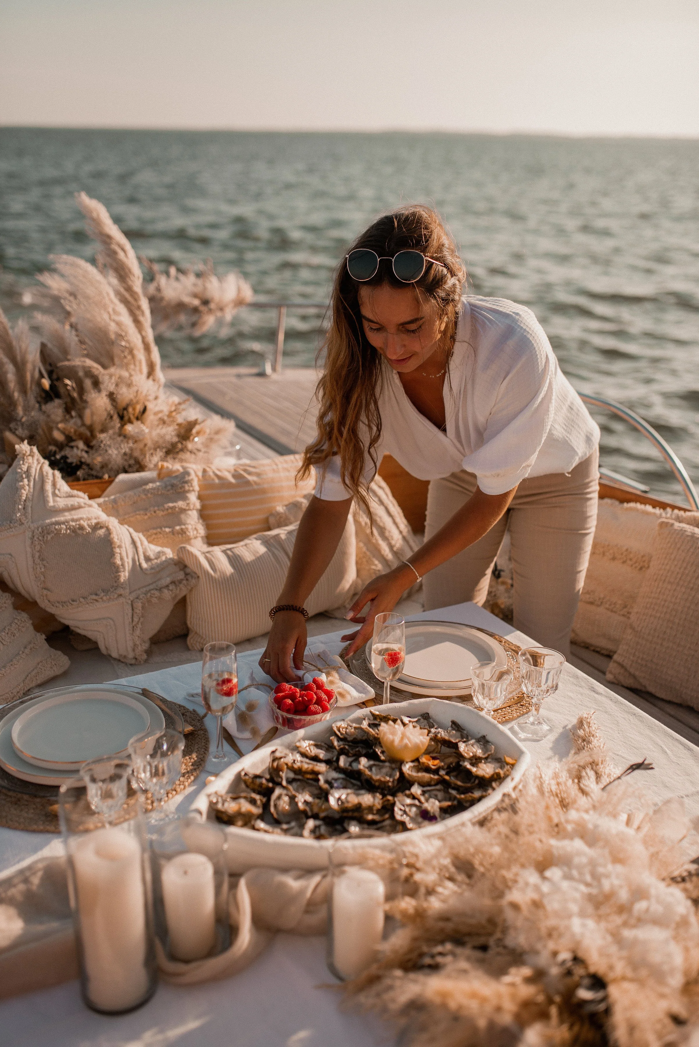 Une femme organise une table élégamment dressée sur un bateau, avec des coquillages, des framboises et des verres à vin près de l'eau, sous un ciel ensoleillé.