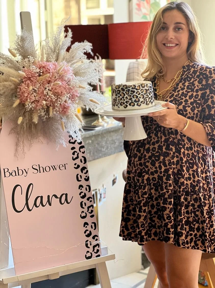 Femme tenant un gâteau à motifs léopard lors d'une baby shower, devant un panneau rose avec "Clara" écrit dessus, décoré de fleurs roses et blanches.