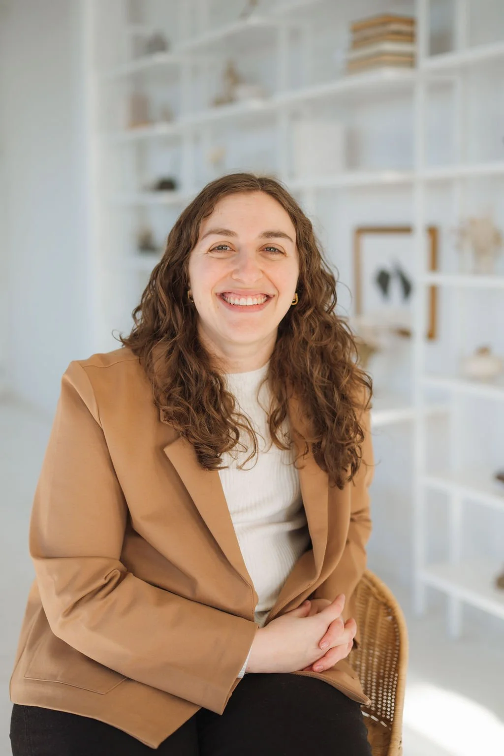 A woman with long, curly brown hair and a big smile, sitting in a modern, well-lit room with white shelves in the background.
