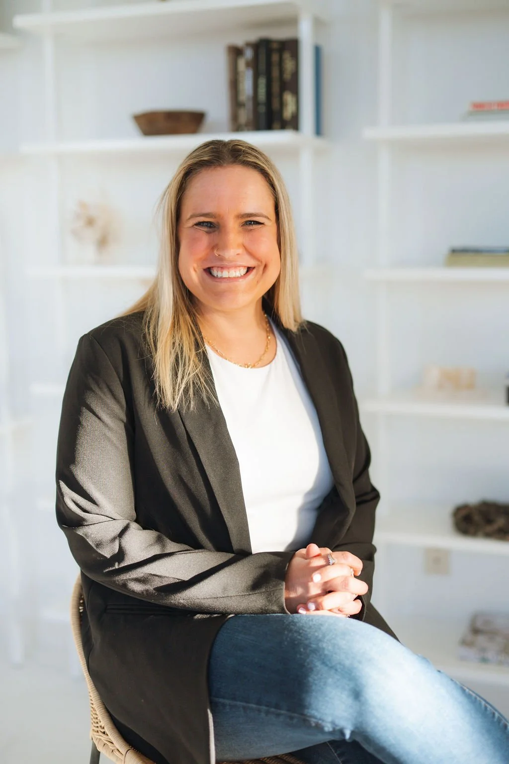 A woman with blonde hair smiling and sitting in a bright room with white shelves in the background.
