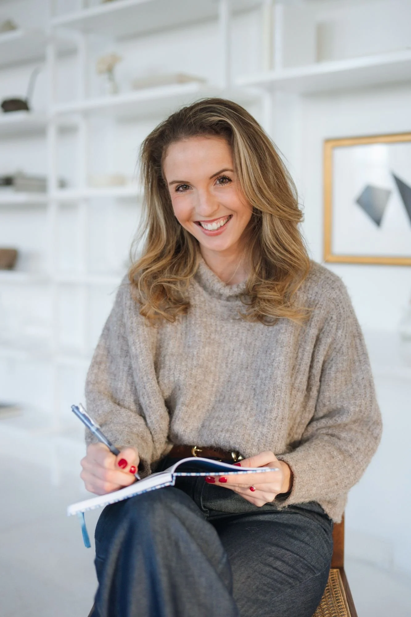 A smiling woman with wavy, shoulder-length blonde hair sitting indoors, holding a notebook and pen, wearing a beige knitted sweater and dark jeans.