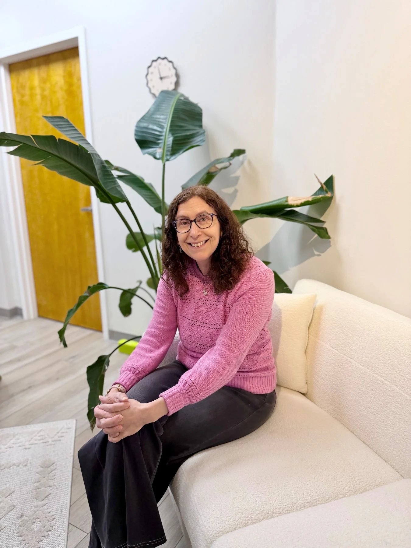 A woman with curly brown hair and glasses sitting on a cream-colored sofa in a room with a large green plant behind her, a yellow door, and a white wall with a clock.