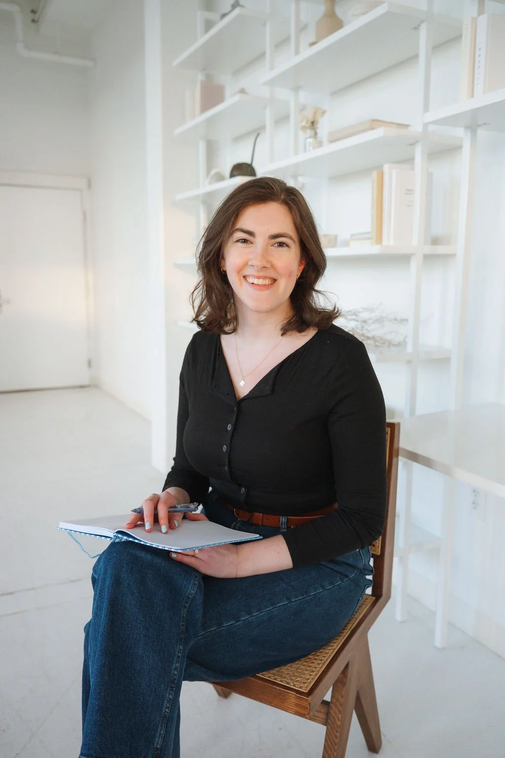 A young woman with shoulder-length brown hair, wearing a black top and blue jeans, is sitting on a wooden chair in a bright room with white decor. She is smiling and holding a notebook and pen.