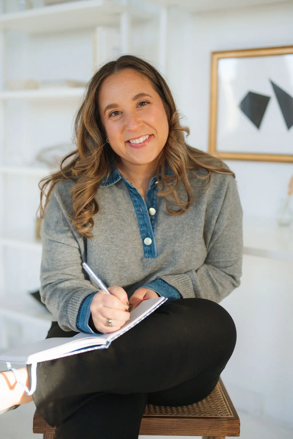 A smiling woman sitting on a wooden chair, holding a pen and notepad, in a bright room with white shelves and a framed abstract art piece in the background.