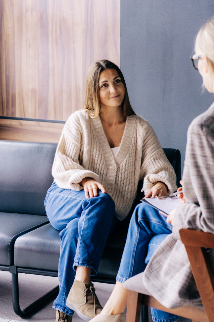 A young woman in a beige cardigan and blue jeans sitting on a sofa, talking to an older woman in glasses holding a notebook in a cozy indoor setting.