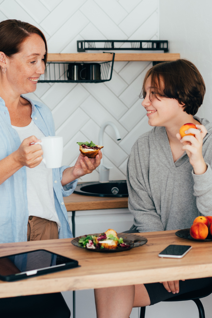 A woman and a teenage boy in a modern kitchen sharing a snack and smiling at each other. The woman holds a mug and a pastry, while the boy has an apple. The kitchen has white tiled walls and wooden countertops.