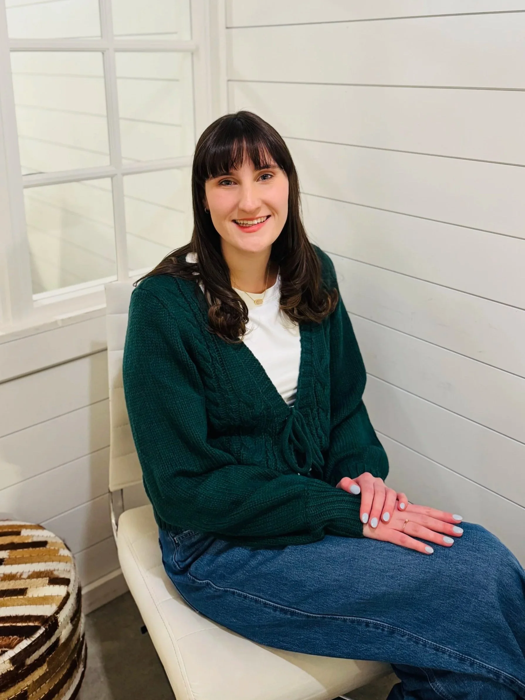 A young woman with long dark hair and bangs, wearing a white shirt, a dark green cardigan, and blue jeans, sitting on a white chair in a room with white paneled walls and a window behind her. She is smiling at the camera with her hands resting on her lap.