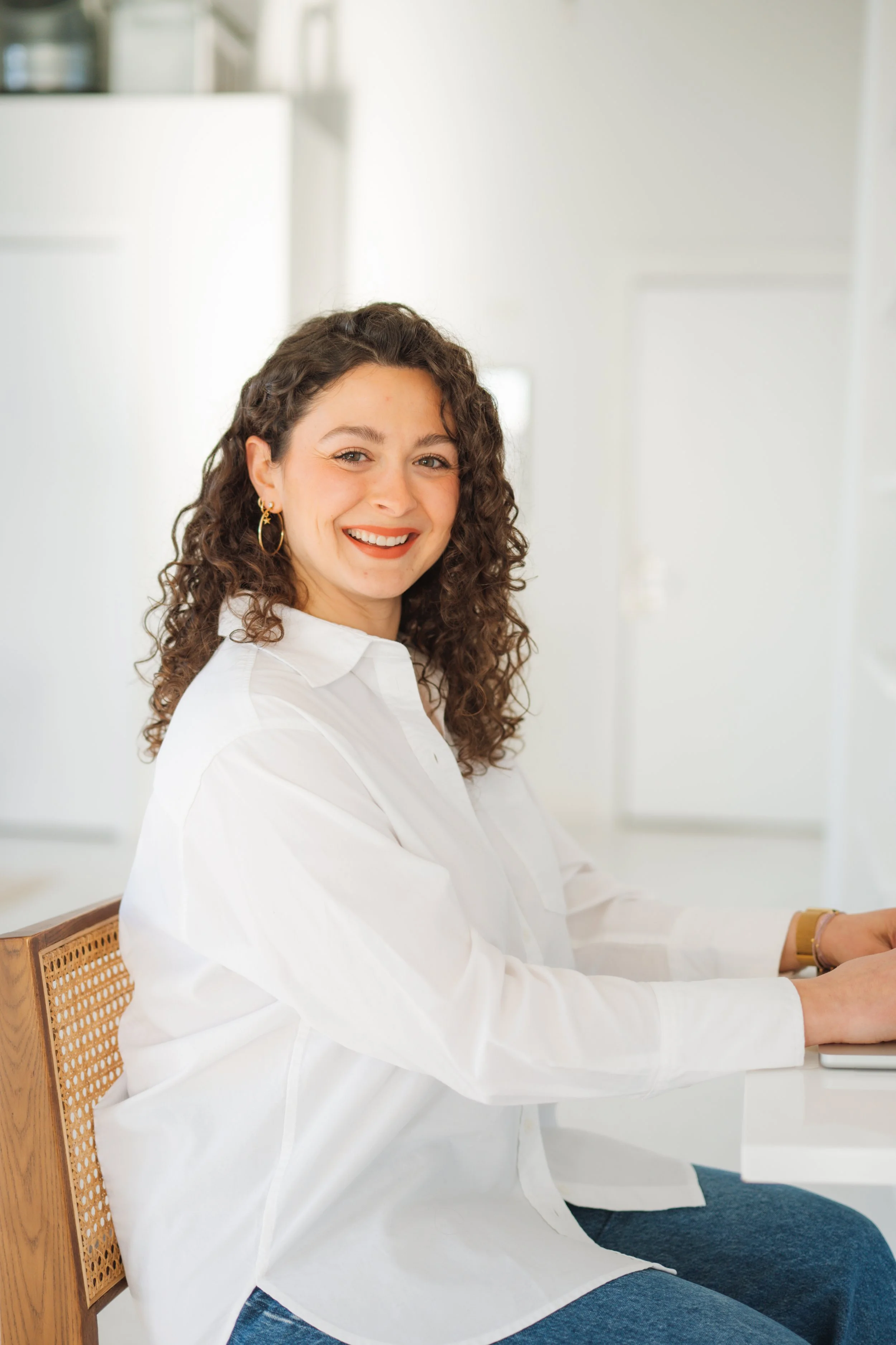 A woman with curly dark hair and a white blouse sitting on a white chair in a modern, minimal office with white walls, a desk with a potted plant, and a closed laptop.