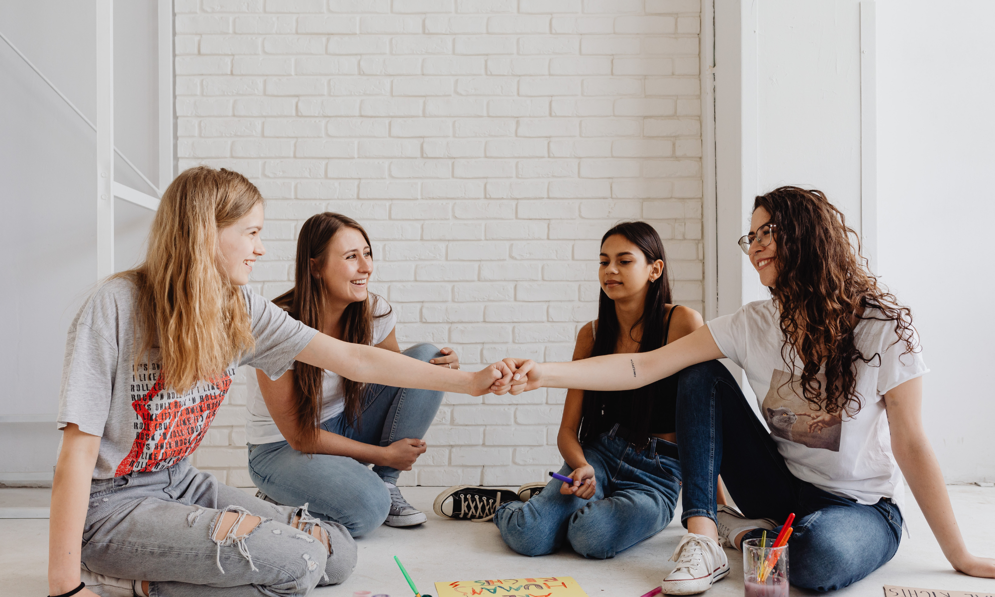Four young women sitting on the floor against a white brick wall, smiling and shaking hands, with art supplies scattered nearby.