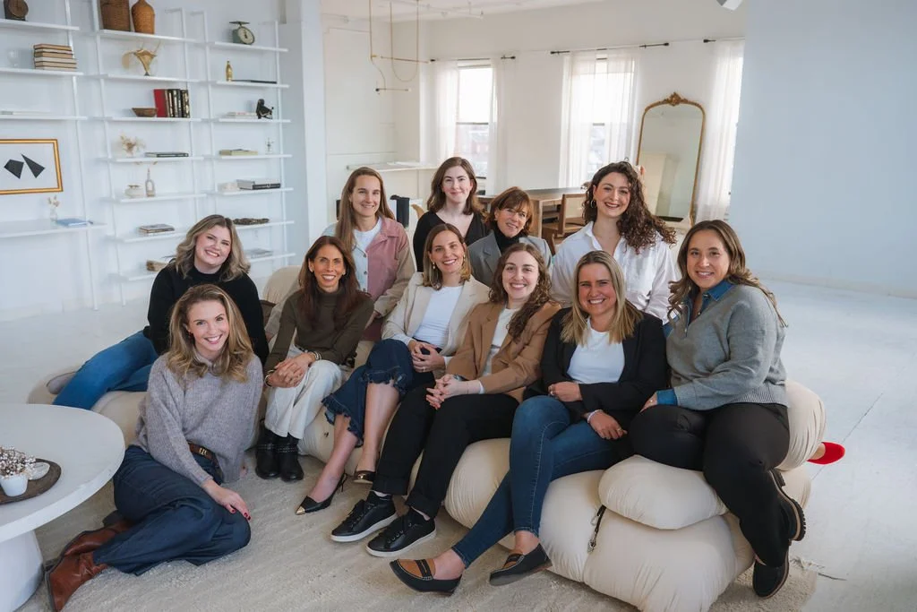 A group of ten women sitting on a white couch in a bright living room with white walls, a large mirror, and shelves with decor and books.