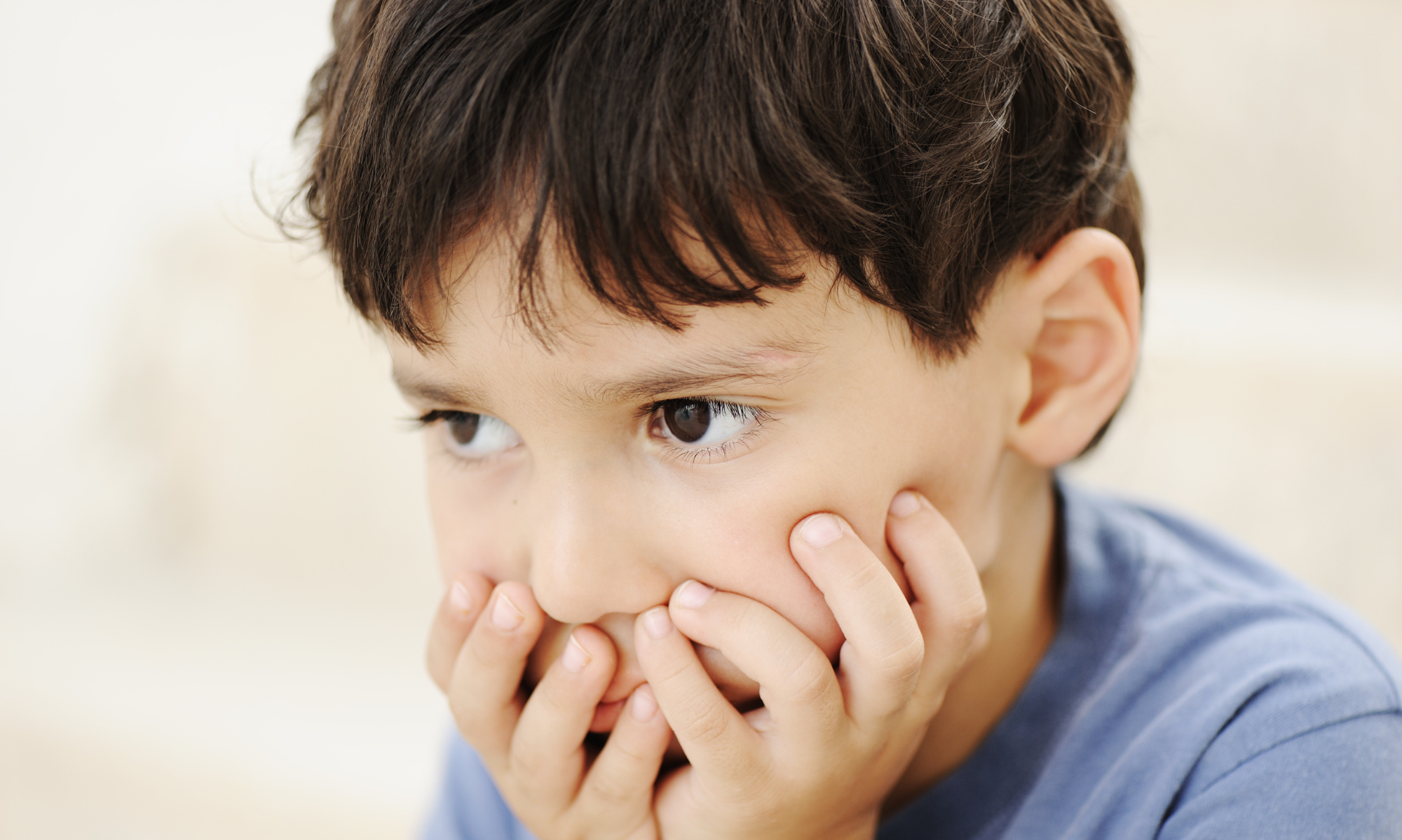 Close-up of a young boy with brown hair covering his mouth with his fingers, looking pensively to the side.