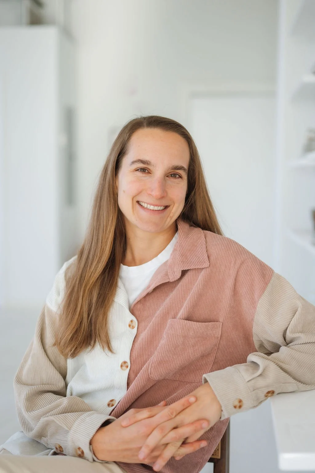 A woman with long brown hair smiling, sitting indoors with a blurred white background, wearing a color-blocked button-up shirt.