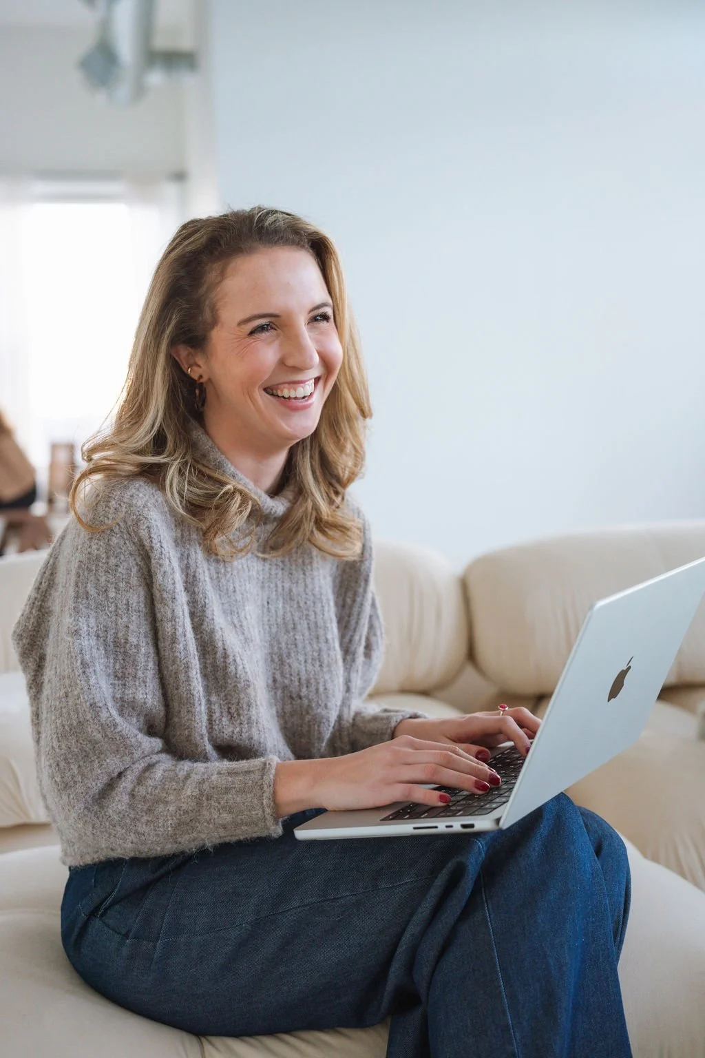 A woman sitting on a beige sofa, smiling and using a silver MacBook laptop.