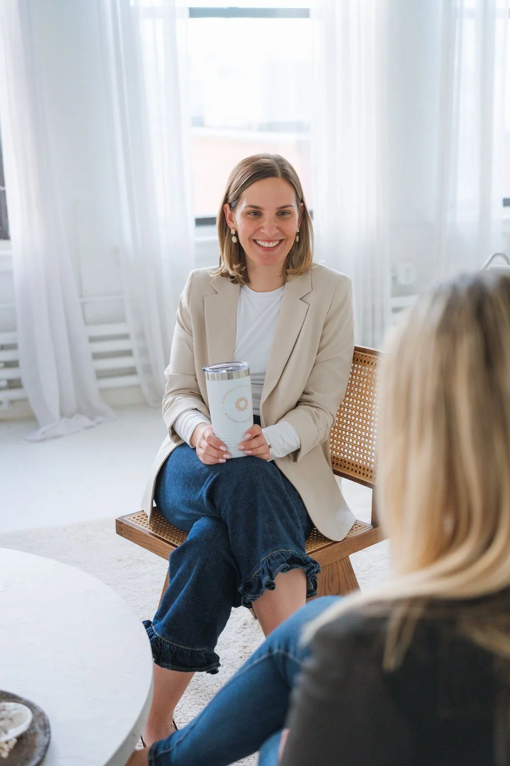 Woman smiling and holding a tumbler while sitting on a chair across from another person in a bright room with white curtains.