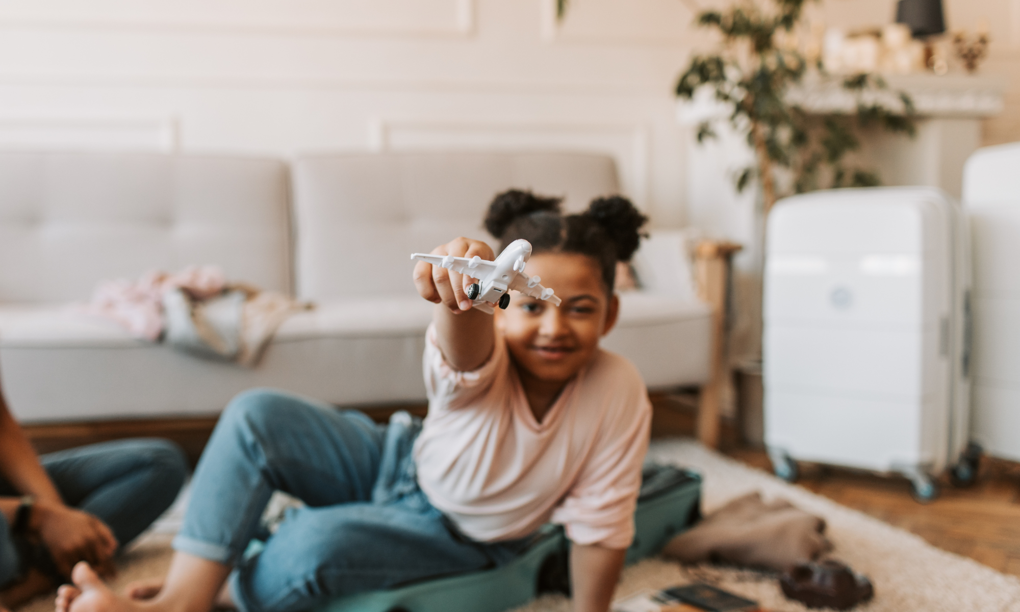 A young girl playing with a toy airplane indoors, sitting on the floor near a suitcase, with a cream-colored sofa and decorative plants in the background.