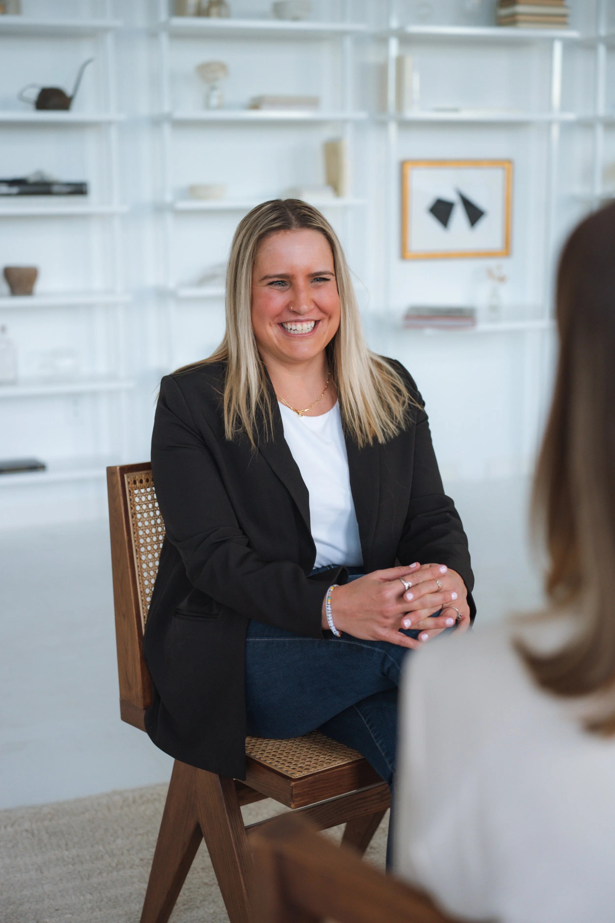 A woman with blonde hair, wearing a black blazer and white shirt, smiling and sitting on a wooden chair in a modern, minimalist room with white shelves and artwork on the wall.