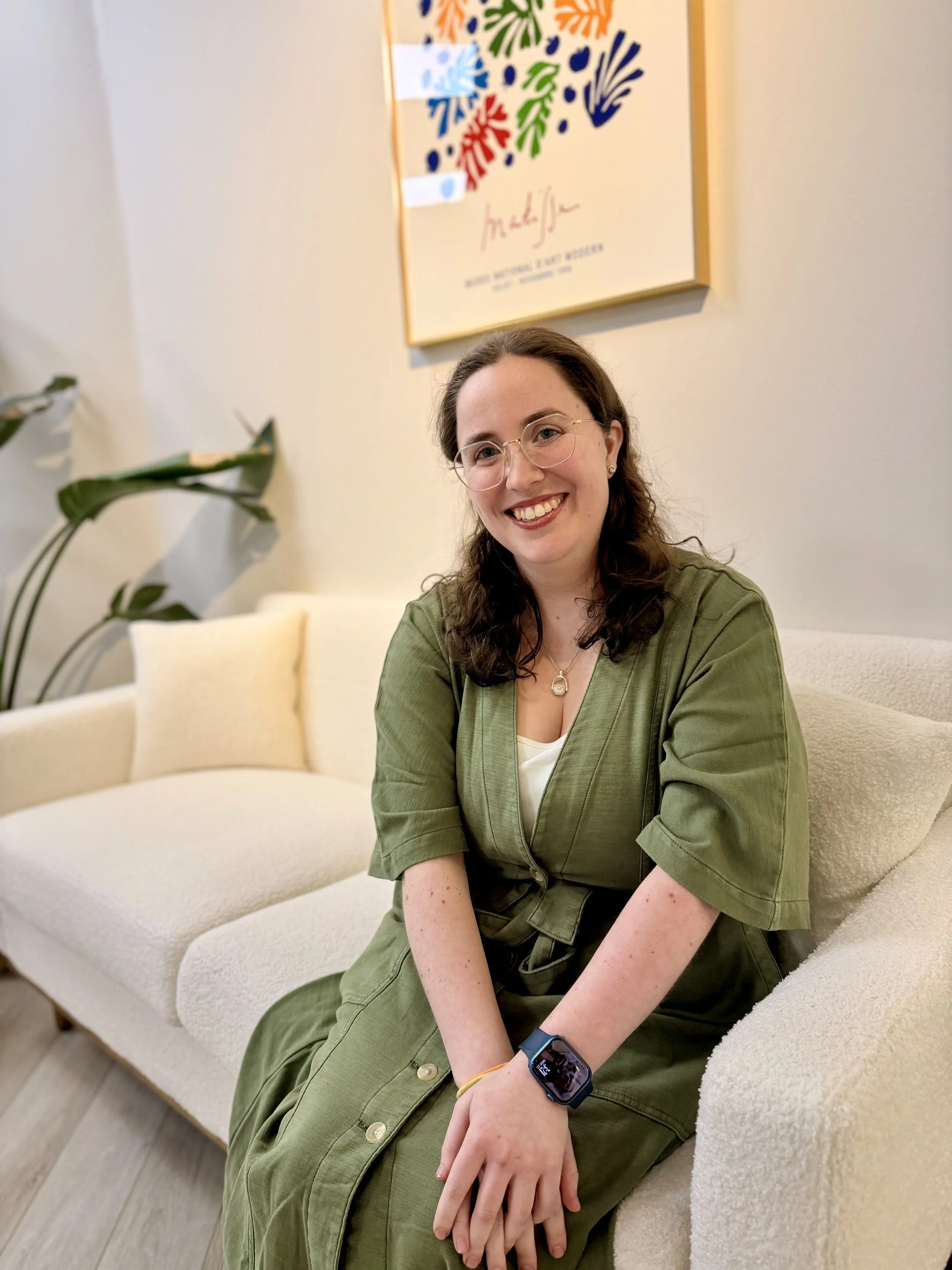 A young woman with glasses and long brown hair sitting on a cream-colored couch in a room with light-colored walls. She is wearing a green dress, smiling at the camera, with a smartwatch on her left wrist and a yellow hair tie on her right wrist. There is a plant on the left and a colorful abstract painting hanging on the wall behind her.