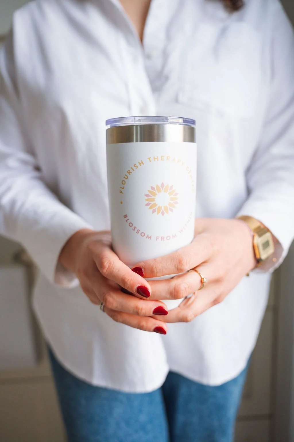 Person holding a white tumbler with a gold-colored lid and a logo that reads 'Flourish Therapy Co.', 'Blossom from Within', and has a pink and orange flower design. The person is wearing a white button-up shirt, blue jeans, a gold watch, and a ring with a small gemstone. Their nails are painted red.