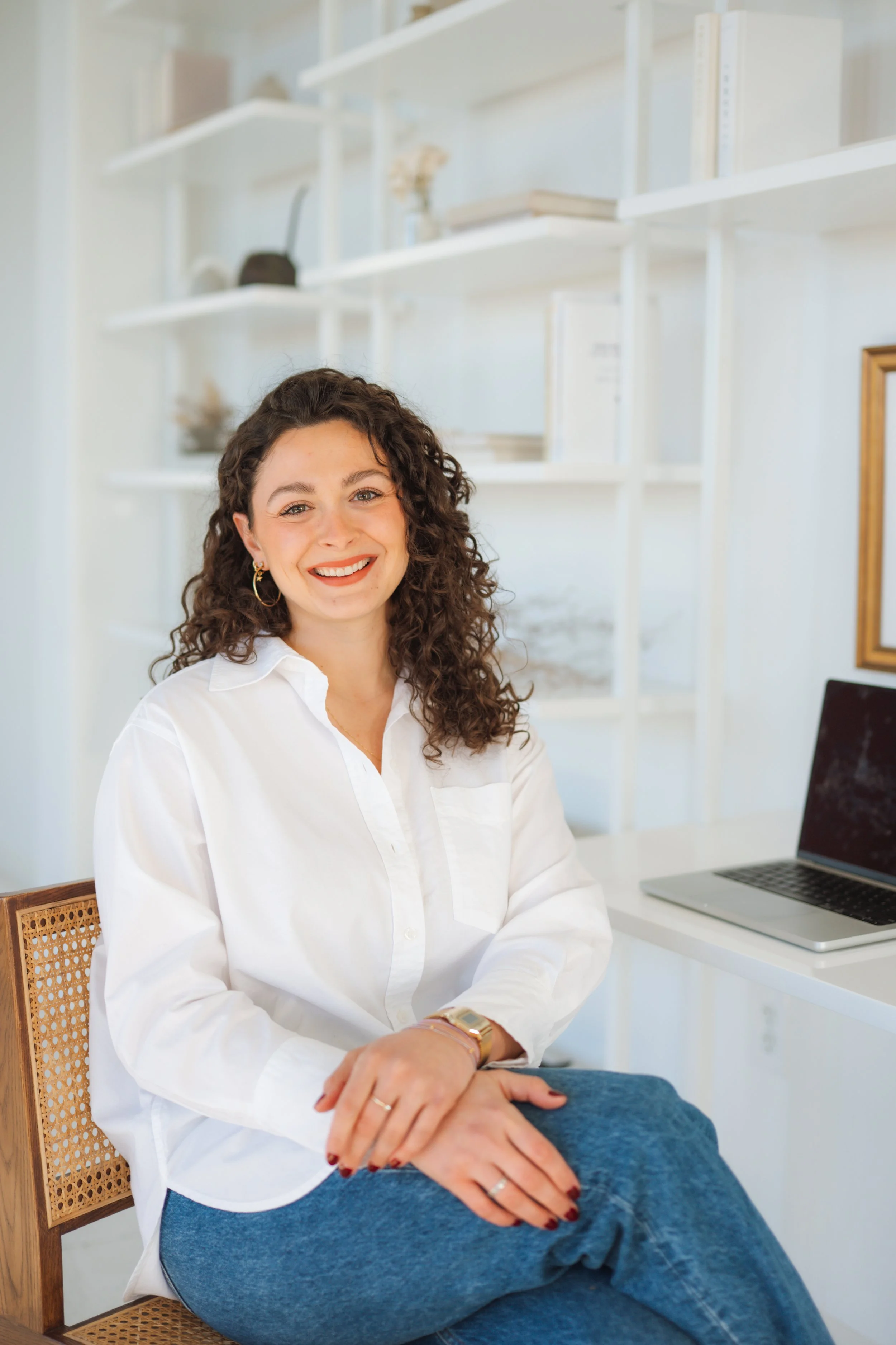A woman with curly dark hair and a white blouse sitting on a white chair in a modern, minimal office with white walls, a desk with a potted plant, and a closed laptop.