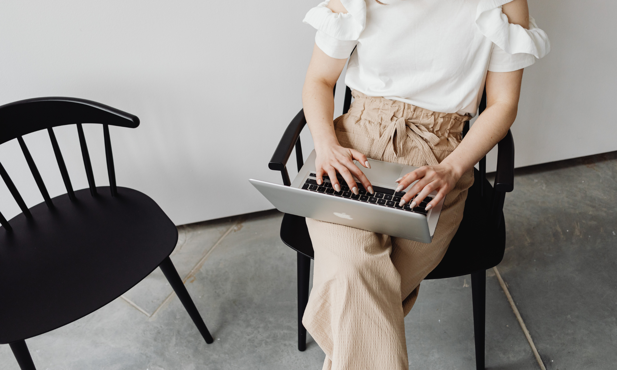 A woman sitting on a black chair with a laptop on her lap, with an empty black chair next to her, on a concrete floor and plain white wall background.