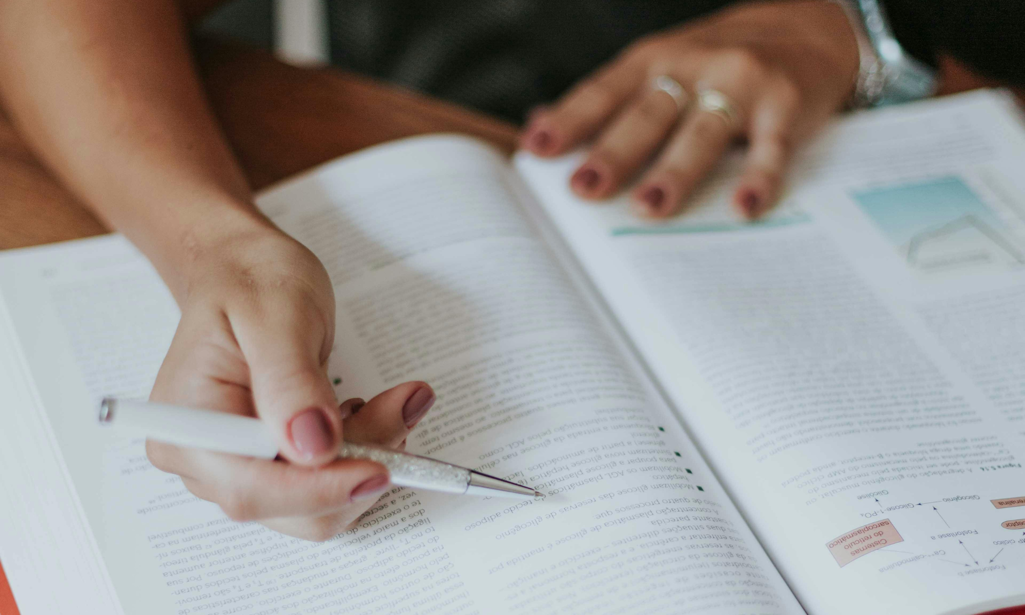 Hands of a person holding a pen and writing in an open textbook on a wooden surface.