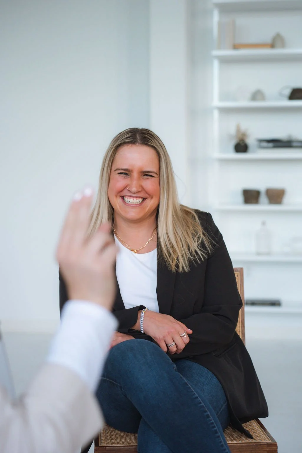 A woman with blonde hair and a black blazer sitting and smiling during a conversation in a well-lit room.