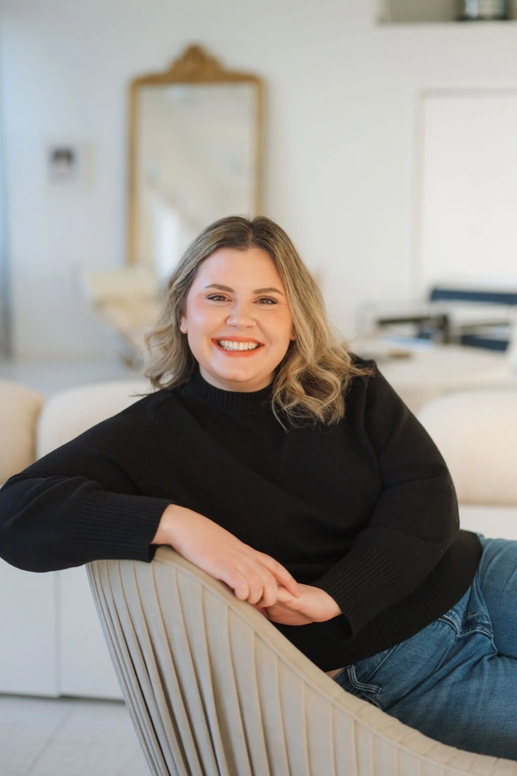A woman with shoulder-length blonde hair and a black sweater, smiling and sitting comfortably in a cream-colored chair in a bright, modern room.