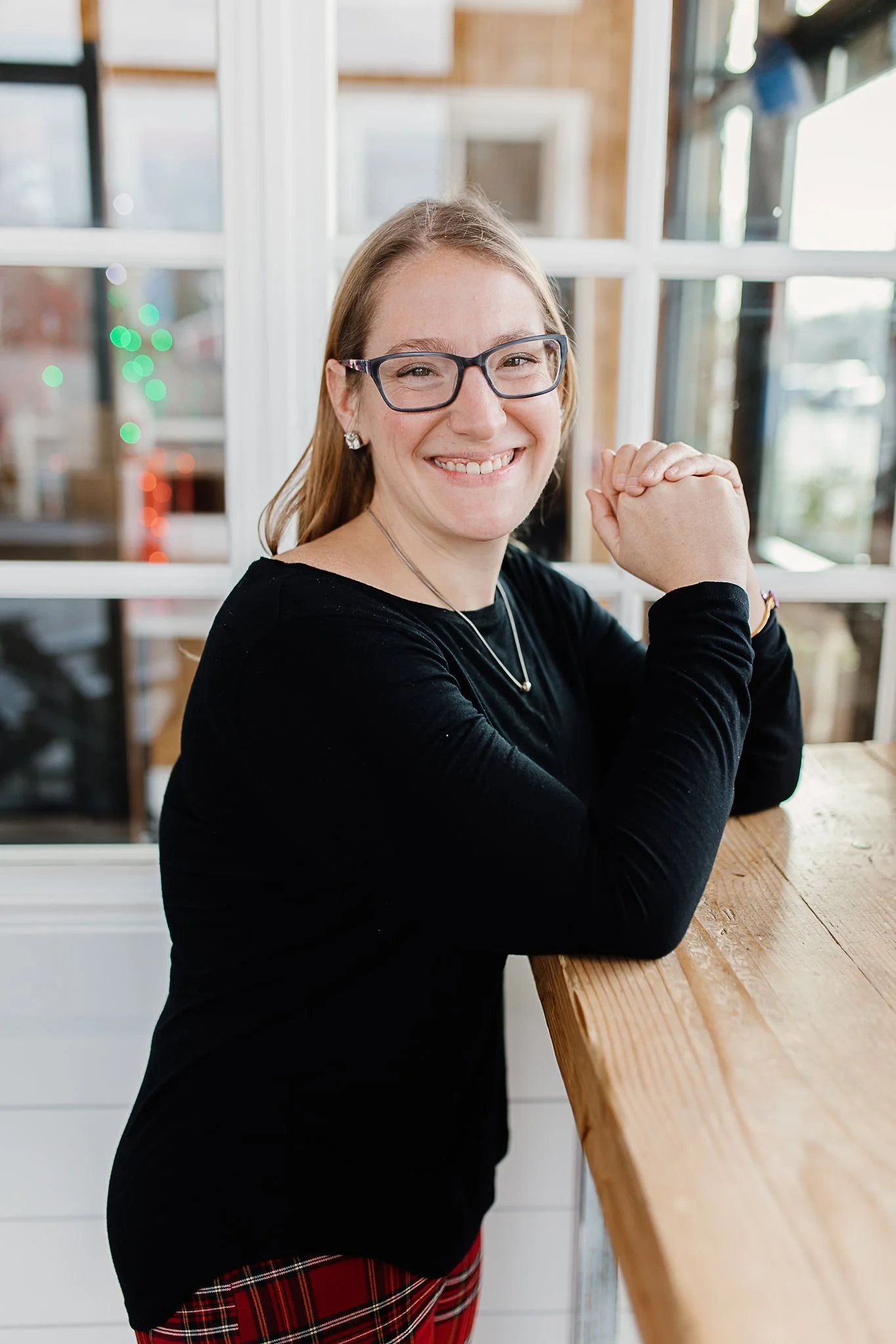 Smiling woman with glasses and earrings sitting by a wooden counter in a bright, modern space.