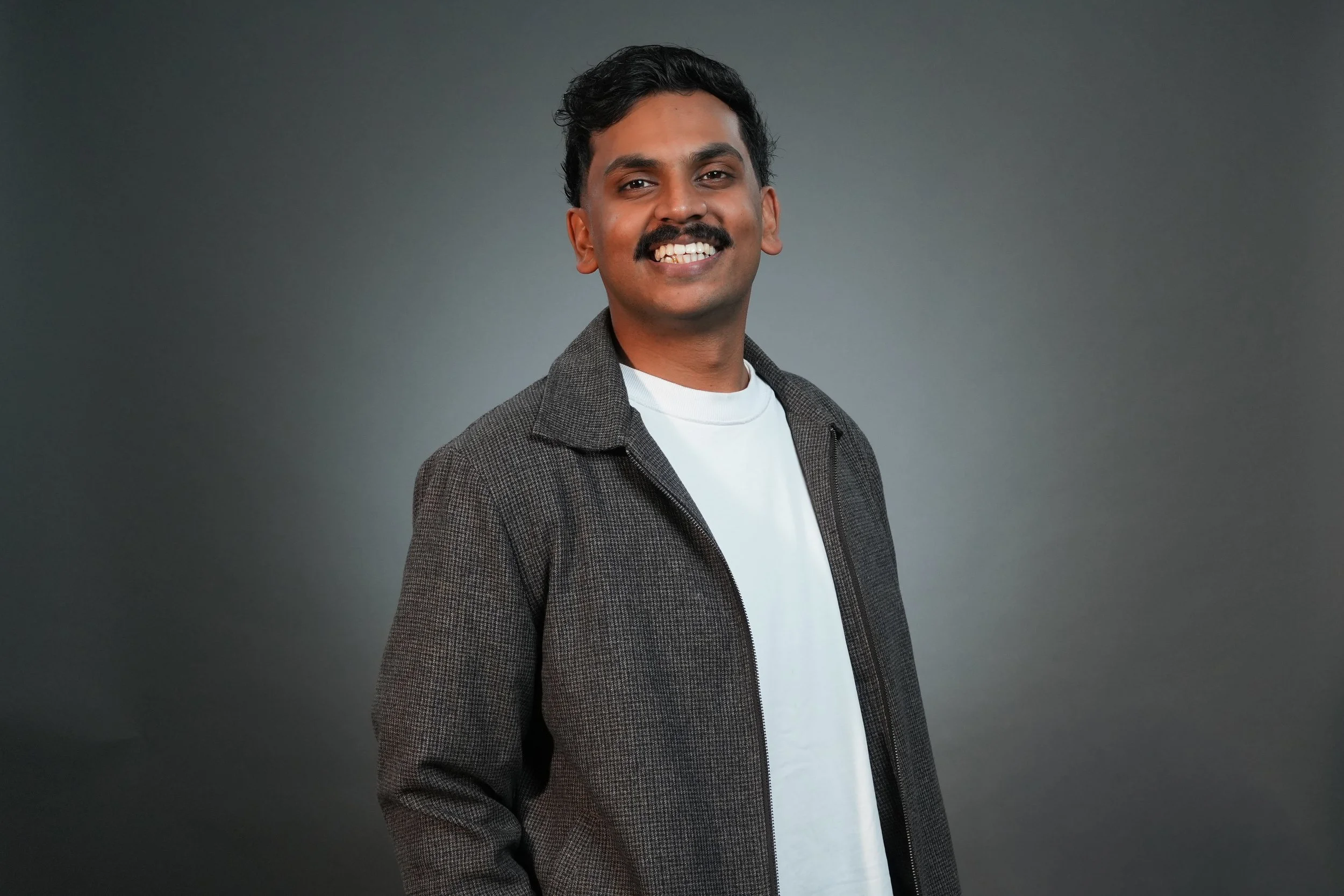 Smiling man in plaid blazer and white shirt with hands in pockets against a gray background.
