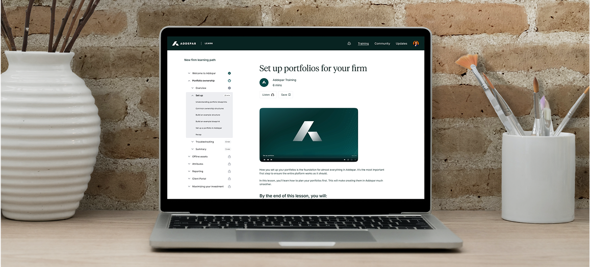 Laptop on a wooden desk displaying an online course titled 'Set up portfolios for your firm' from Adddepar. The desk has a white vase with brown sticks on the left and a white container with paintbrushes on the right, against a brick wall background.
