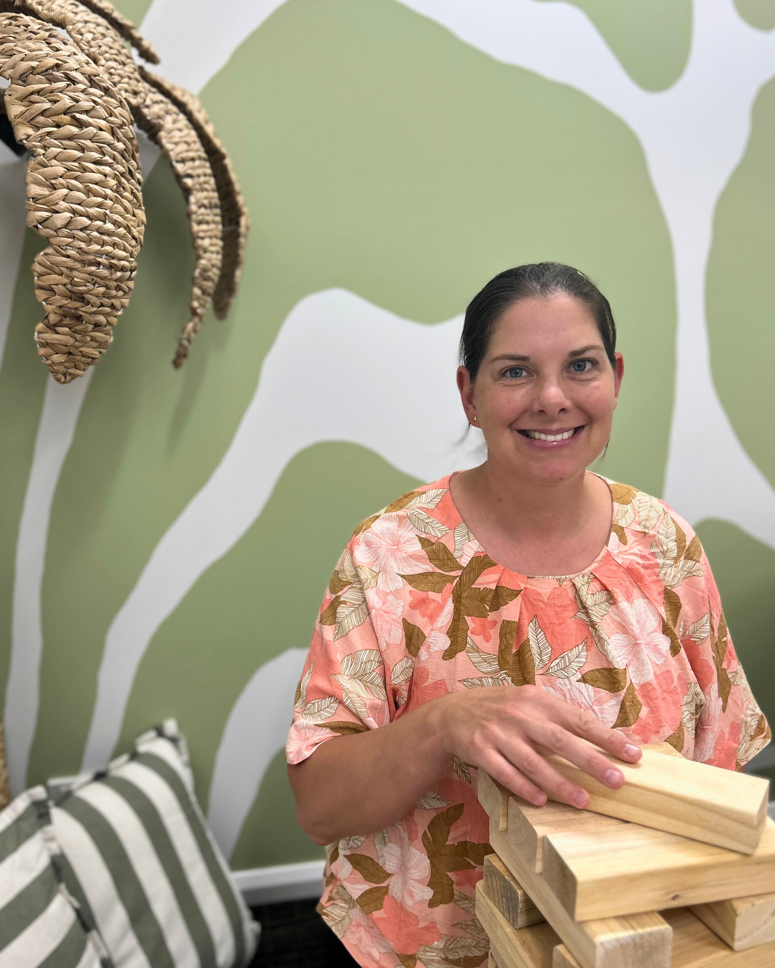 A woman with dark hair smiling, wearing a floral pink and brown shirt, standing in front of a green and white abstract patterned wall, holding wooden blocks in her hands.