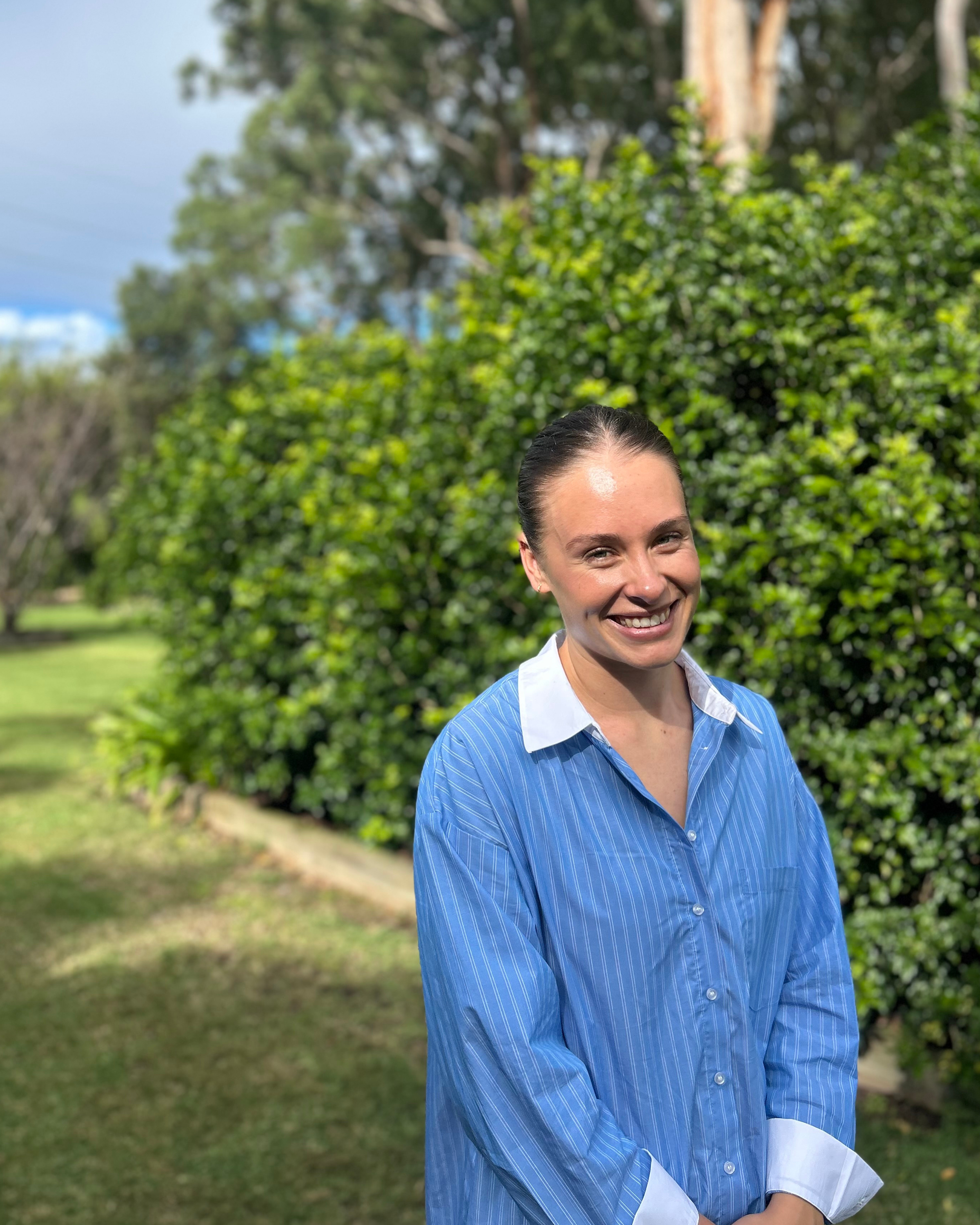 A woman with wet hair wearing a blue striped shirt smiling outdoors with green bushes and trees in the background.