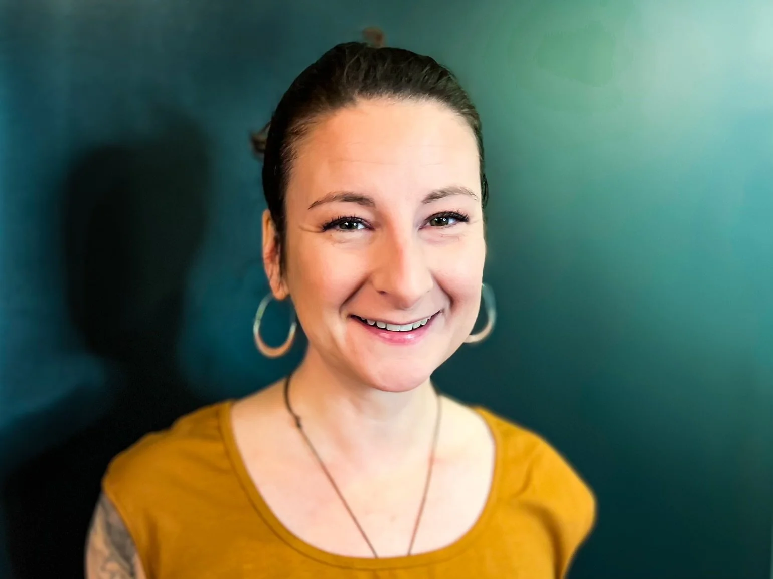 An image showing the artist, Katie, agains a black backdrop from shoulders up. She is smiling, has brown hair, pulled back into a bun, is wearing silver hoop earrings, and an ochre colored short sleeve top.