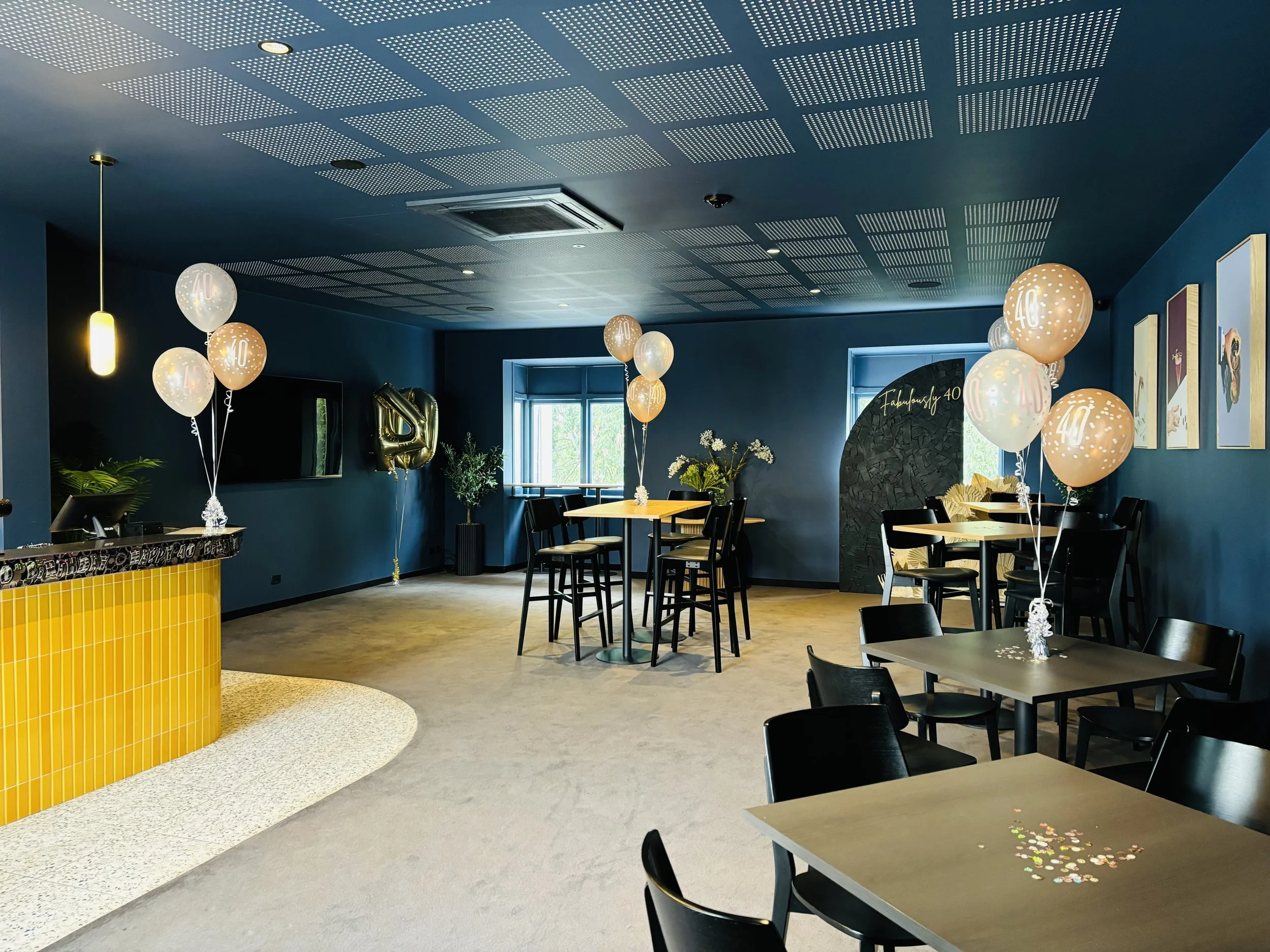 A decorated celebration room with blue walls, black tables and chairs, balloons with '40' labels, and a yellow-tiled counter, indicating a 40th birthday party.
