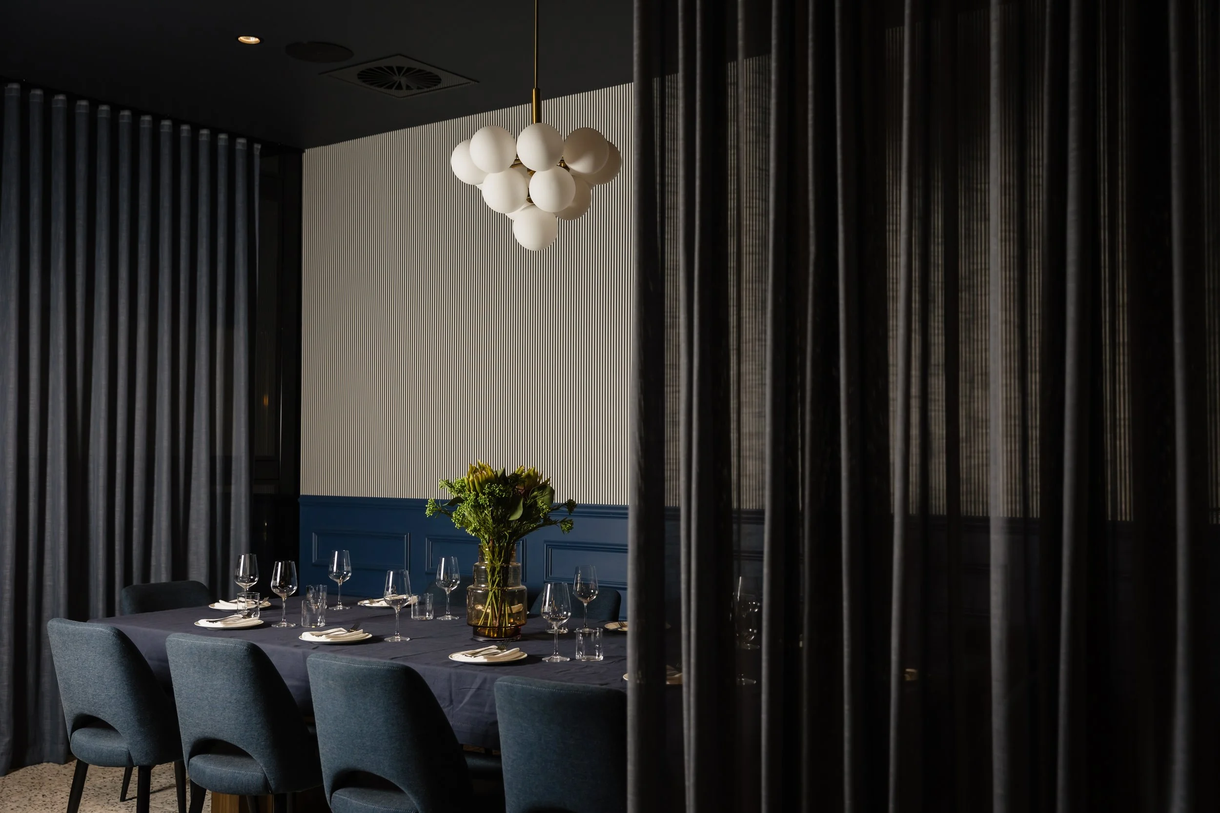 Elegant dining room with a table set with wine glasses and plates, a large centerpiece with flowers, dark curtains, and a modern white chandelier hanging from the ceiling.