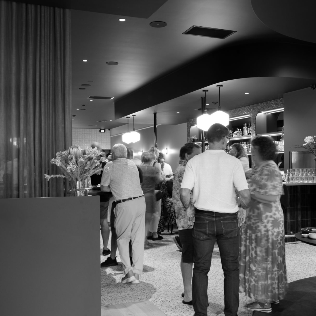 People socializing in a bar with a bar counter, shelves of liquor bottles, and hanging lights.