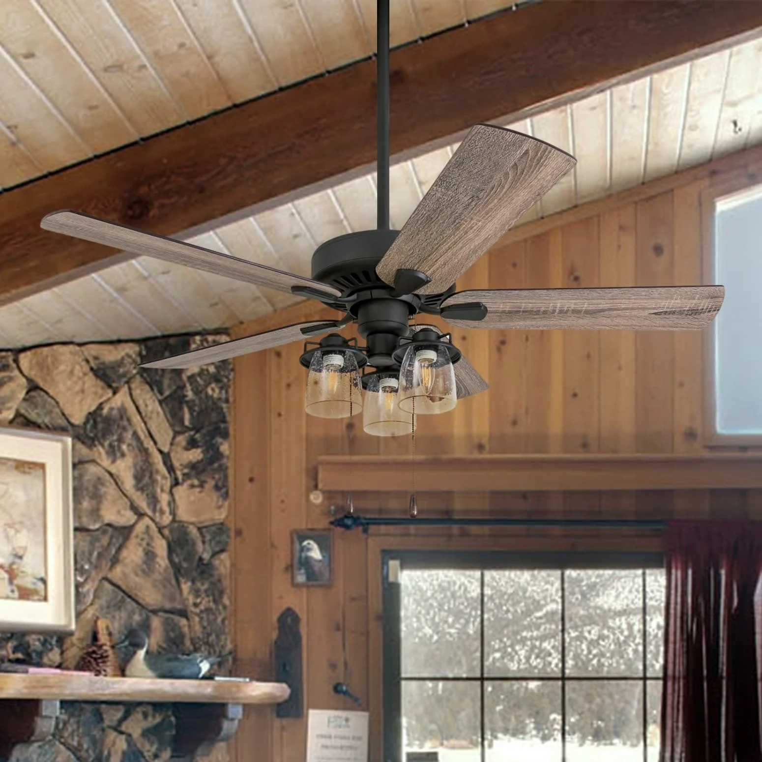 Rustic wooden ceiling with modern ceiling fan and glass light fixtures in a cozy log cabin interior with stone fireplace and snowy window view.
