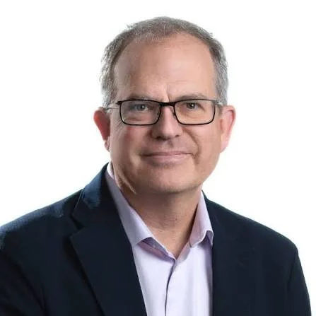 A man with grey hair smiles at the camera. He is light-skinned and wears black glasses, a light purple button up, and a navy blue blazer.