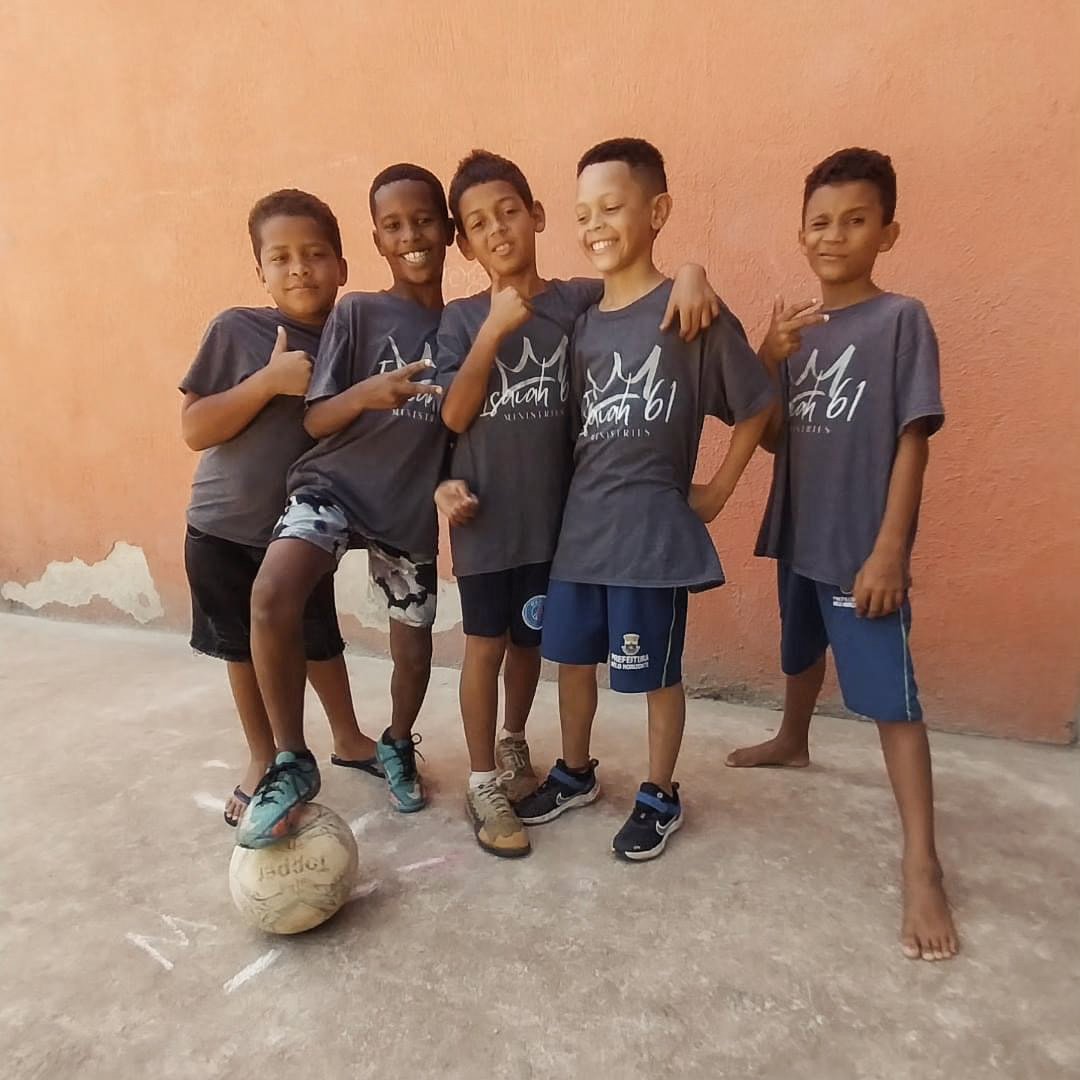 Five young boys standing together outdoors, wearing matching dark T-shirts, with a soccer ball on the ground near them against an orange wall.