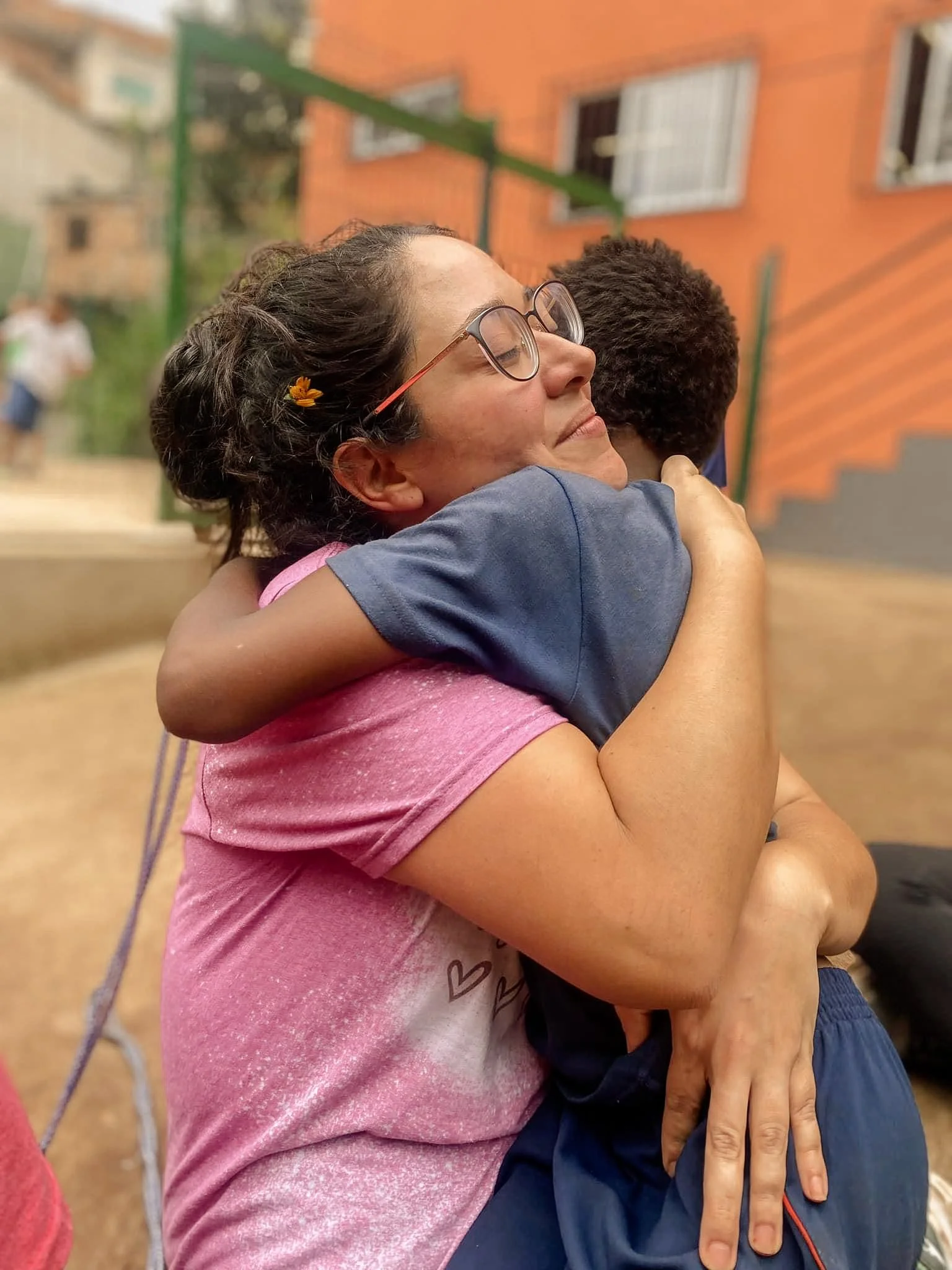 A woman with glasses and a pink shirt hugging a child with curly hair and a blue shirt outdoors, with a brick building in the background.