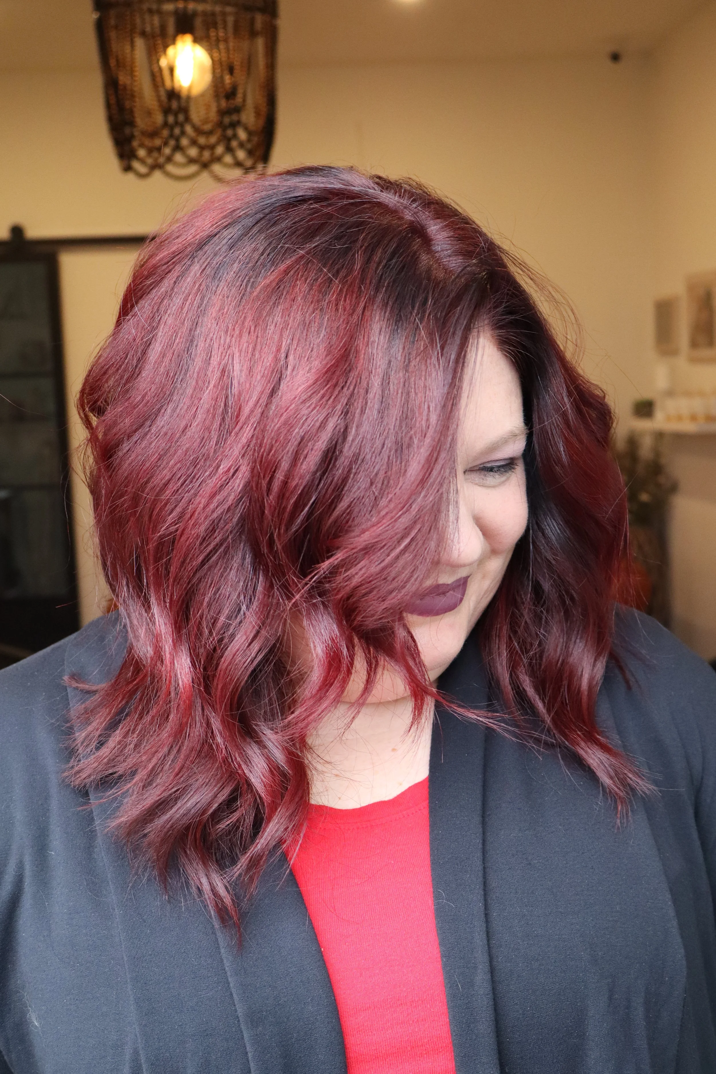 Close-up of a woman with shoulder-length wavy hair dyed in shades of red and dark brown, wearing a red top and black blazer, smiling with her eyes closed.