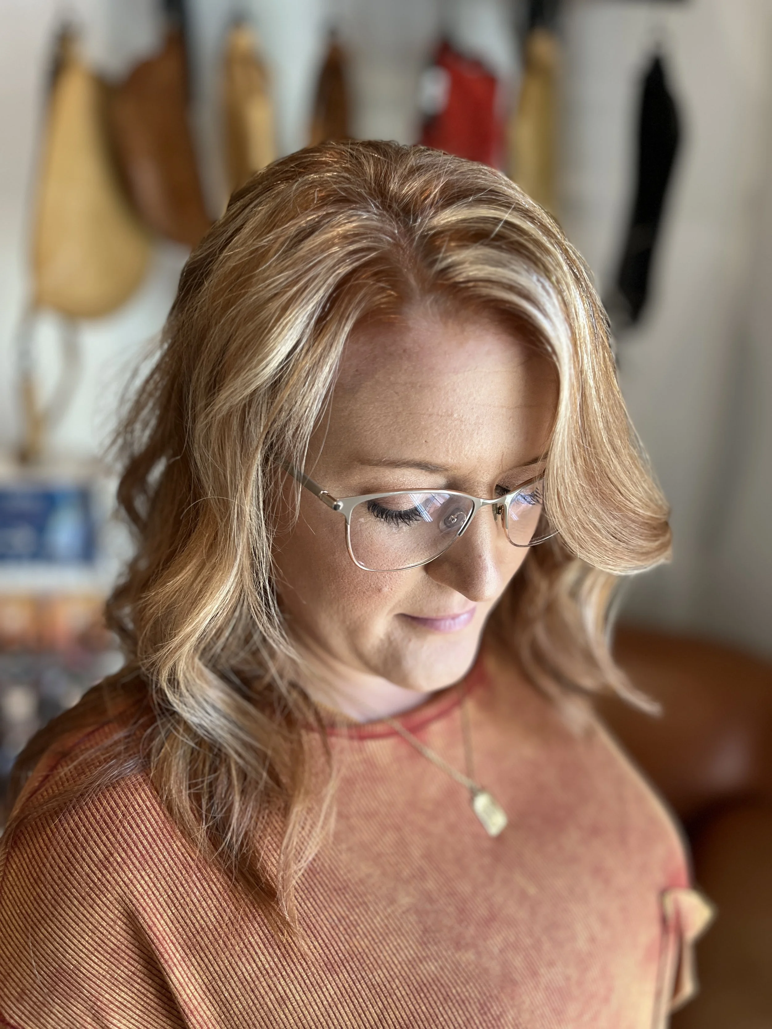 Close-up of a woman with blond hair, wearing glasses and a rust-colored top, looking down with a slight smile. In the blurred background, various handbags are hanging on a wall.