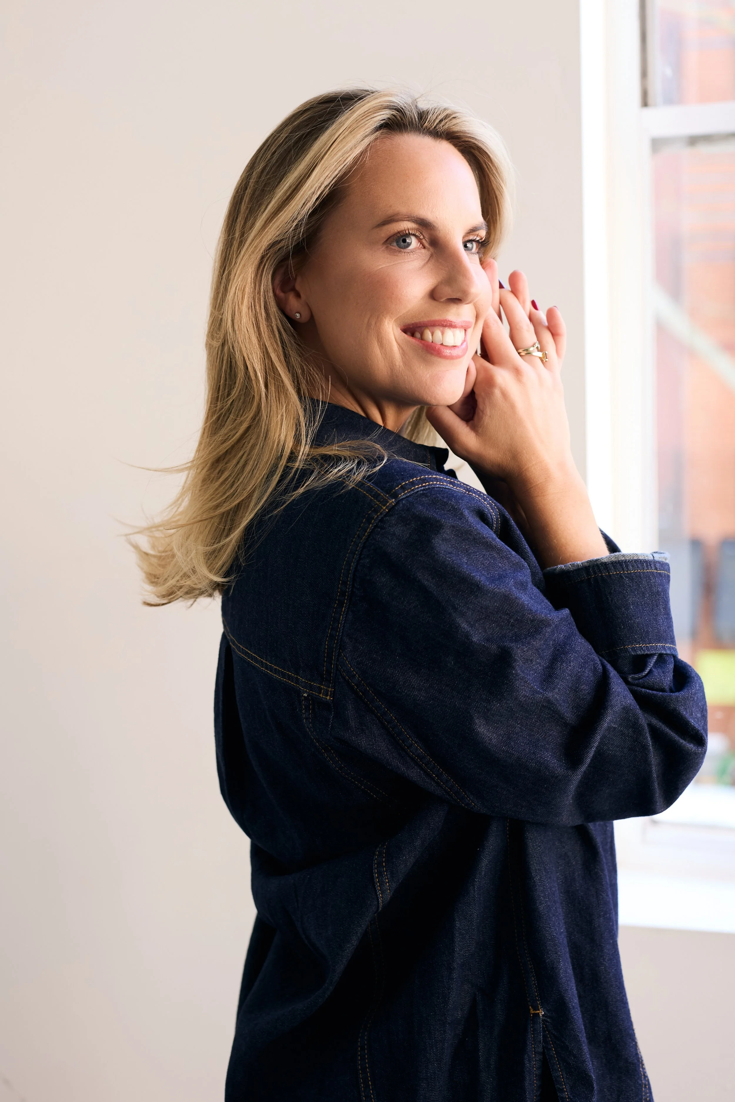 A woman with blonde hair smiling, wearing a dark denim jacket, near a window with natural light.