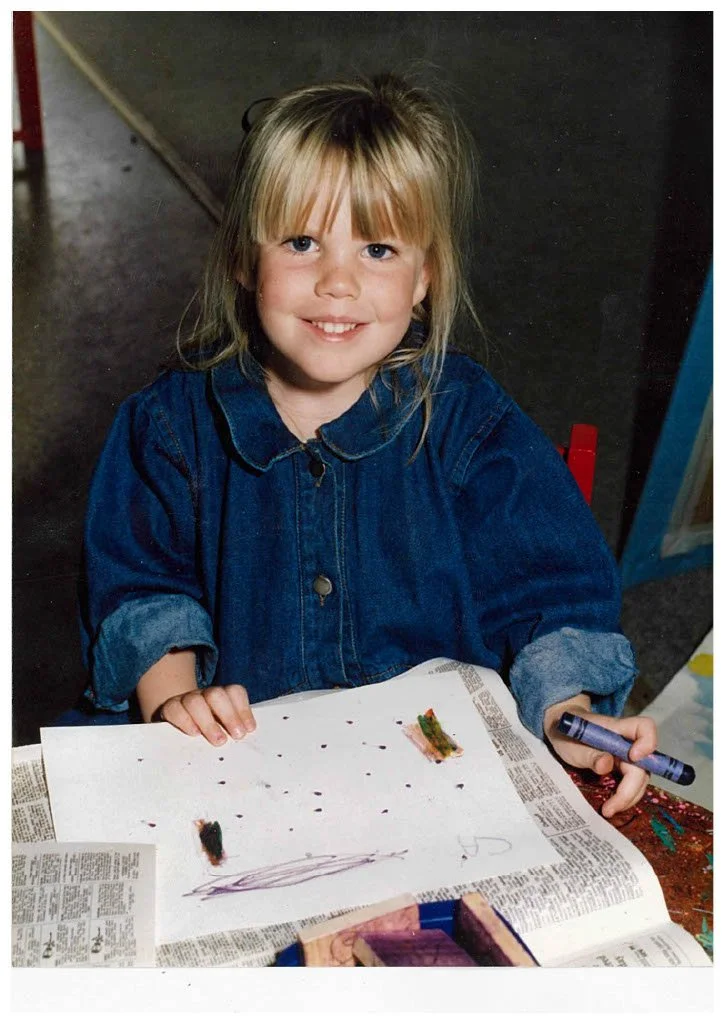 A young girl with blonde hair and a big smile sits at a table with a marker in her hand, showing her artwork on a piece of paper.