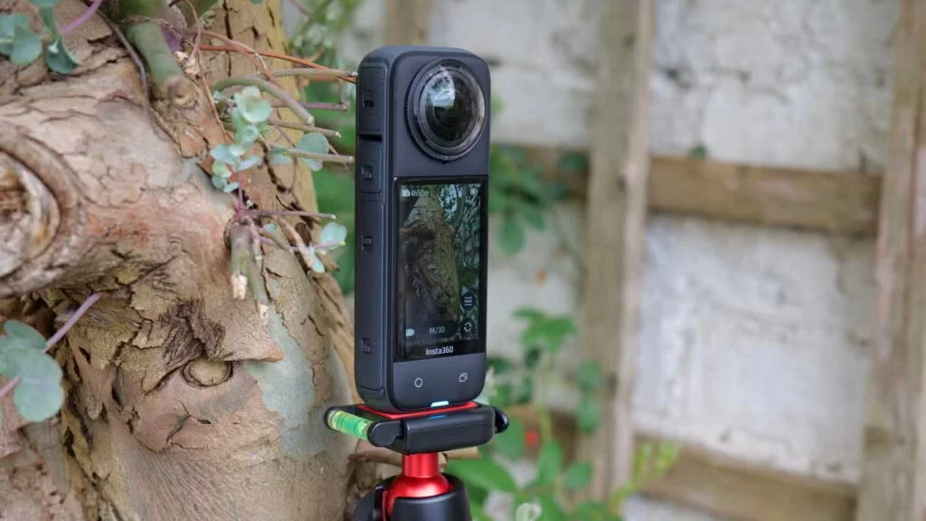 An Insta360 X3 mounted on a small tripod leaning against a tree trunk in an outdoor garden setting.