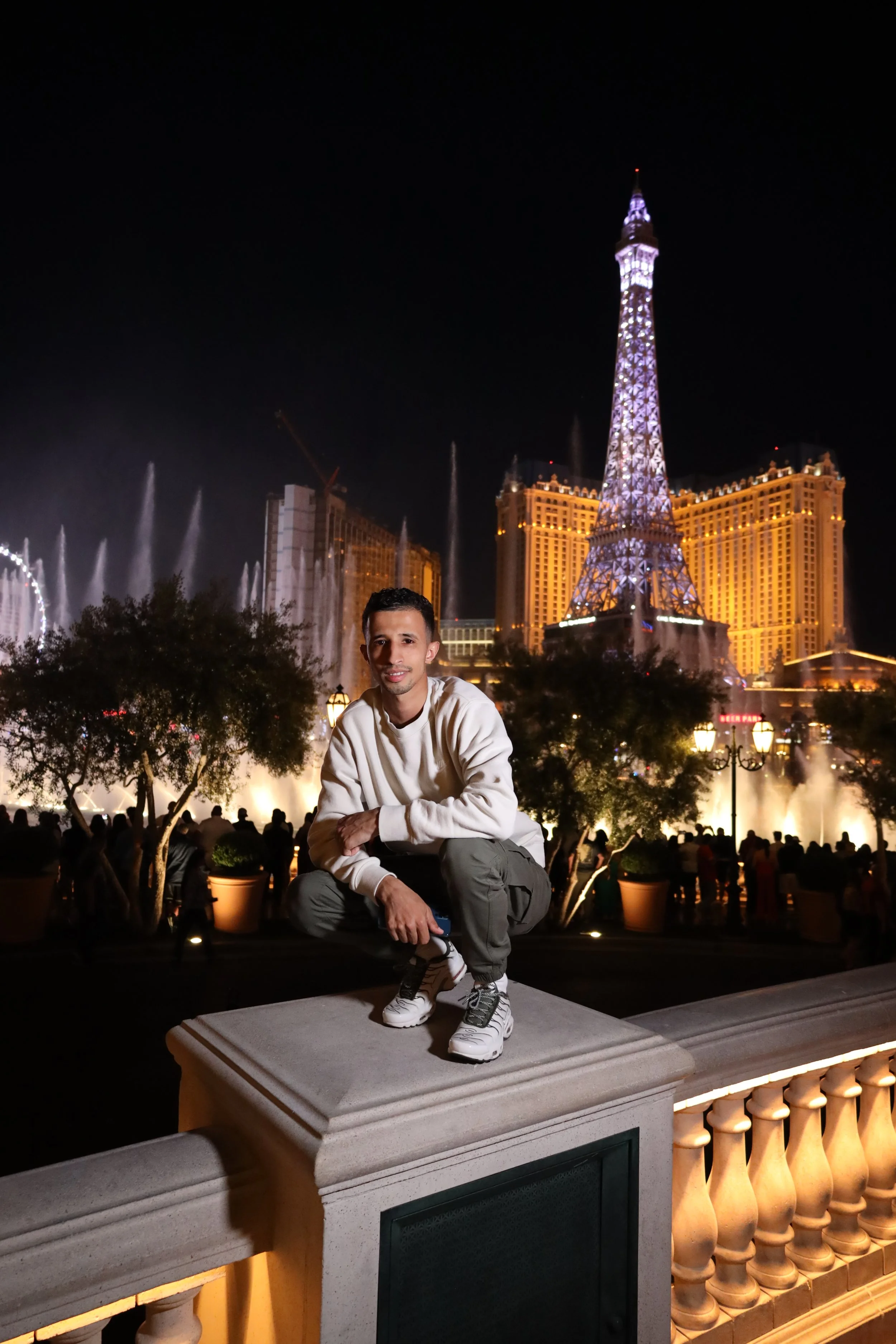 A man crouching on a balcony with the Las Vegas Strip at night behind him, featuring the Eiffel Tower replica and illuminated buildings.
