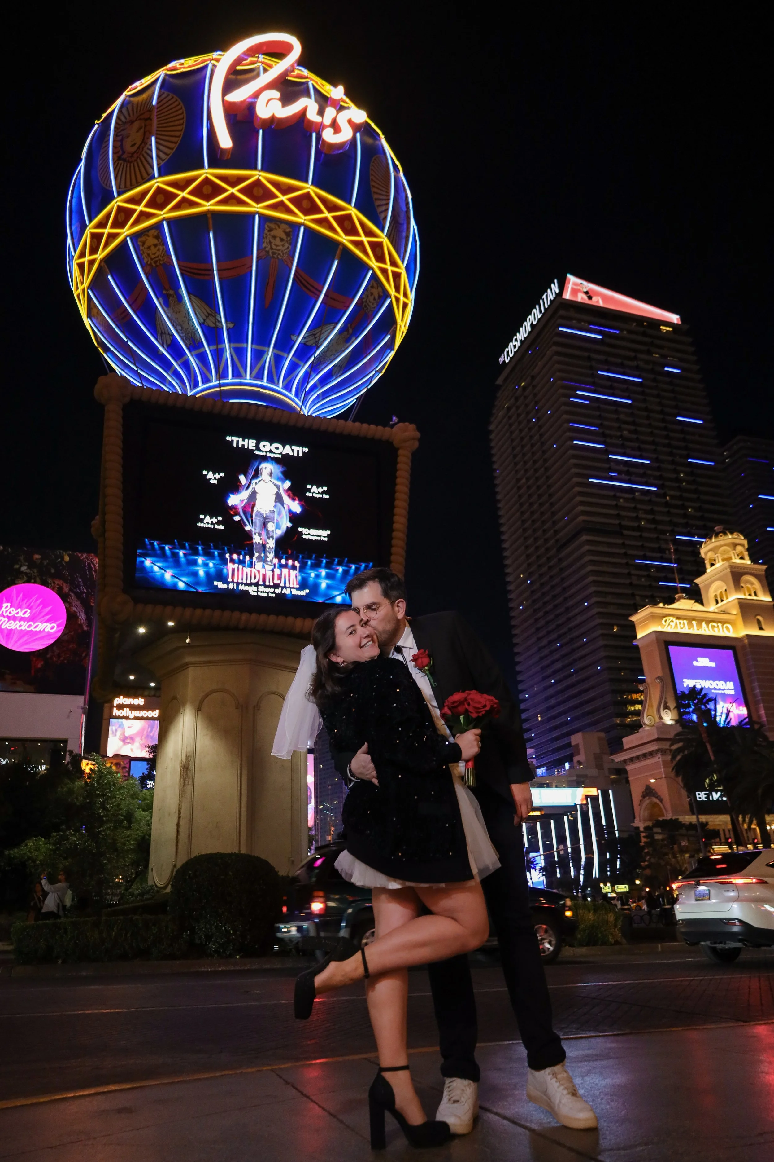 A couple in wedding attire stands kissing on the street at night in front of a large illuminated Paris sign and neon-lit buildings, with bright city lights and vehicles in the background.