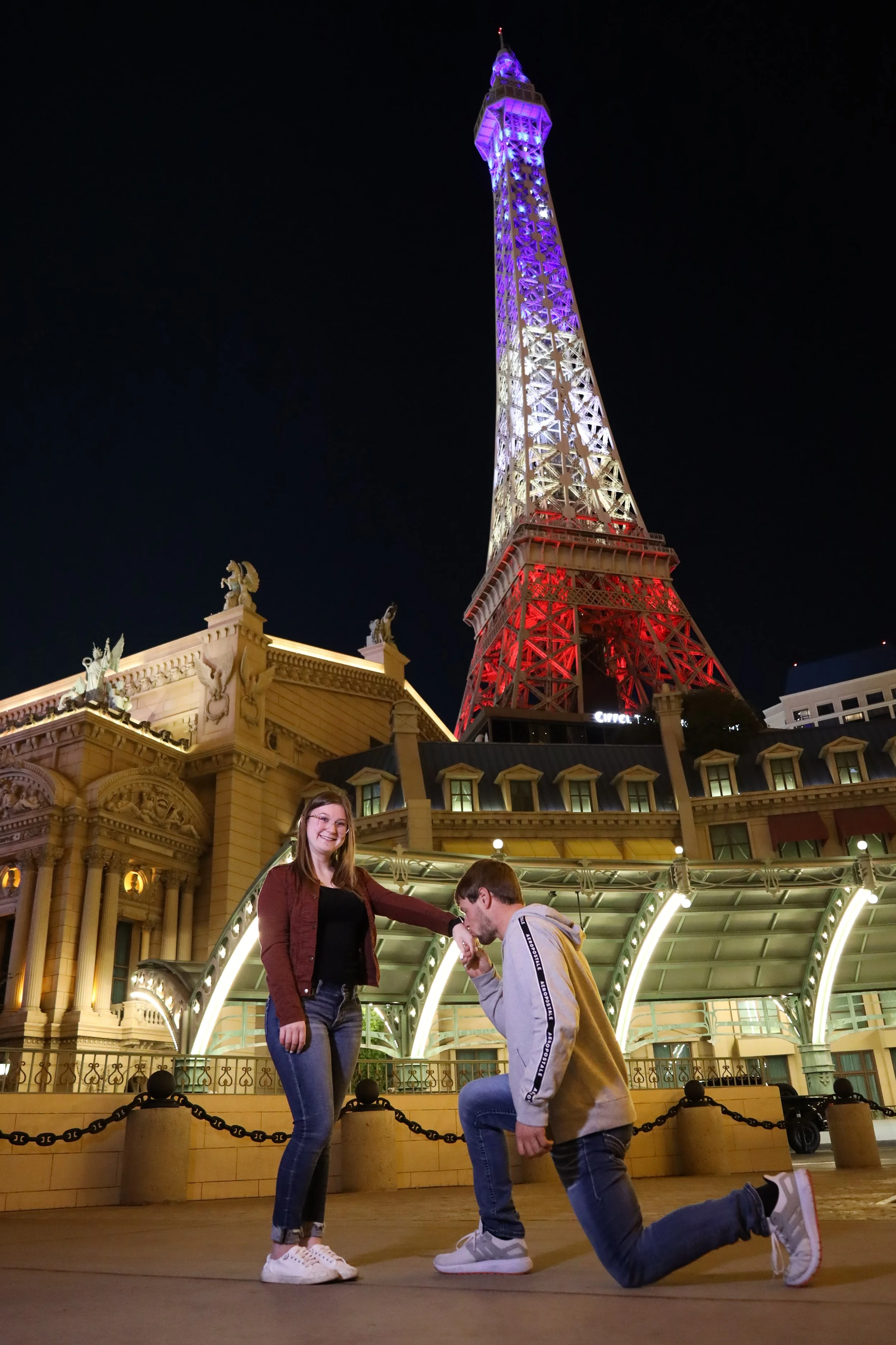A man proposing to a woman in front of the illuminated Las Vegas Eiffel Tower replica at night, with a historic building and city lights in the background.