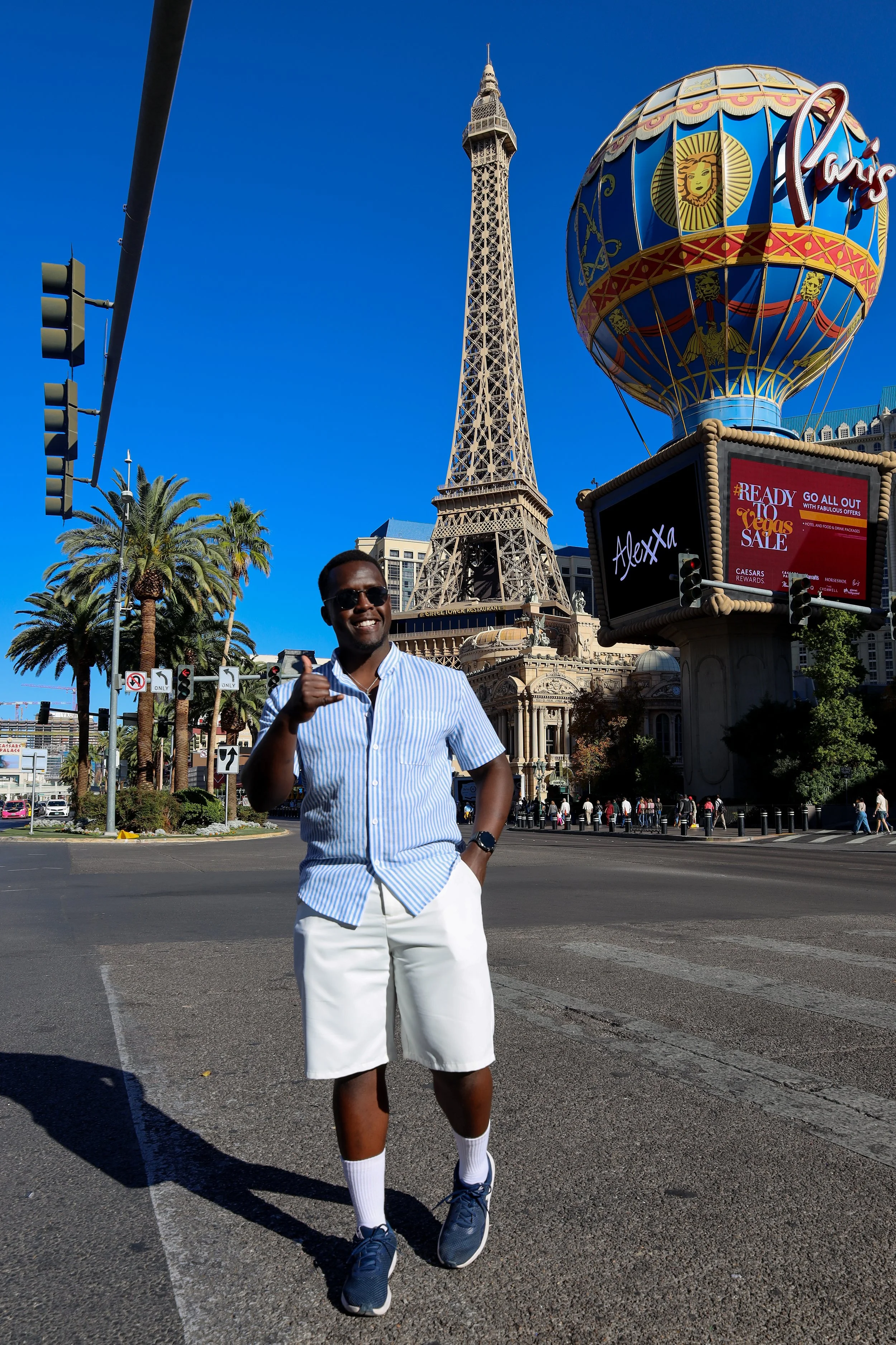 A man in a blue and white striped shirt, white shorts, and sneakers standing on a street with the Paris Las Vegas Hotel replica Eiffel Tower and a hot air balloon sign in the background.
